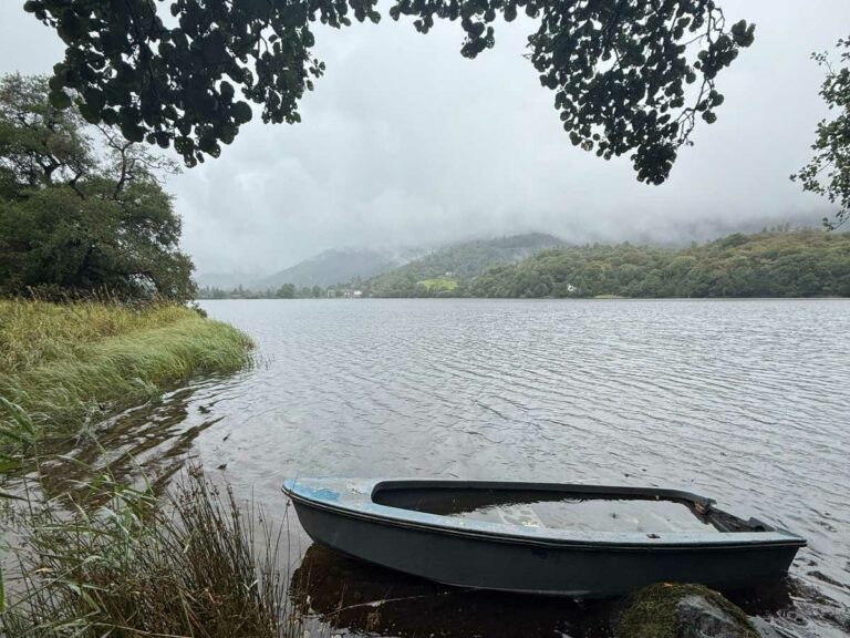 Moored boat on calm lakeshore with forested mountains and low cloud in background