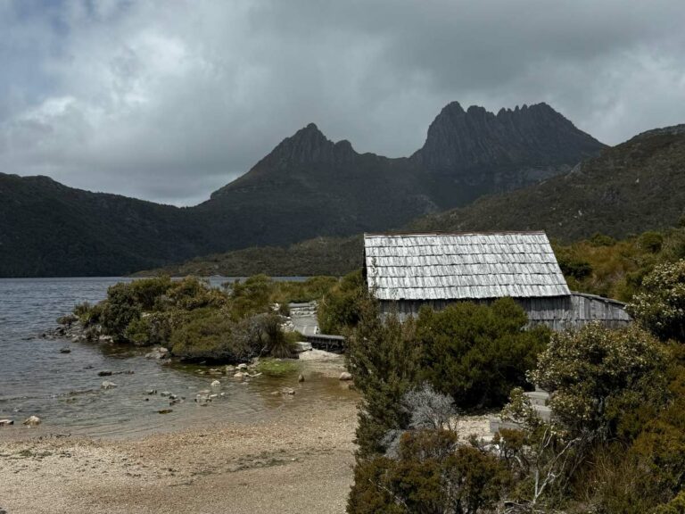 Historic boathouse on sandy beach at Dove Lake with jagged mountain peaks and native Australian bushland