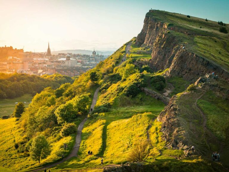 Walking trails lead up to a rocky peak at sunset overlooking a city in the background.