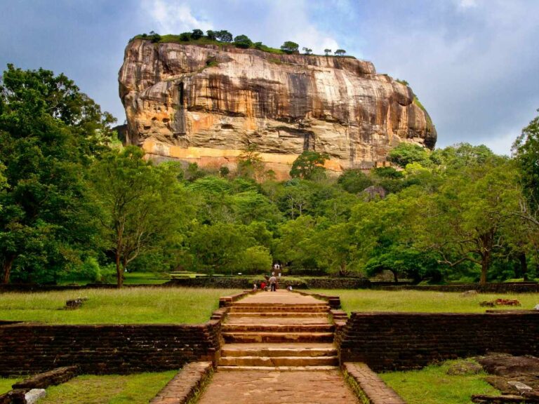 The ancient stone and brick staircase leading up the vertical cliffs of Sigiriya Rock (Lion Rock) in Sri Lanka. Further along, the narrow path is surrounded by lush tropical greenery and ancient fortifications, showcasing the UNESCO World Heritage site's dramatic height and archaeological detail.