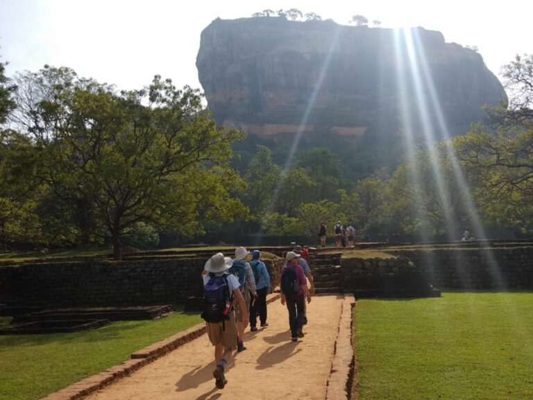 Sun shines behind large rock outcropping as travellers walk up path to stairs leading to climb the rock.