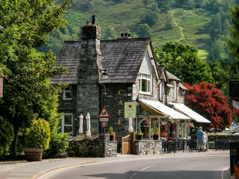 Quaint Village Cafe with stone chimney, flower baskets, tables and chairs and hills in the background.