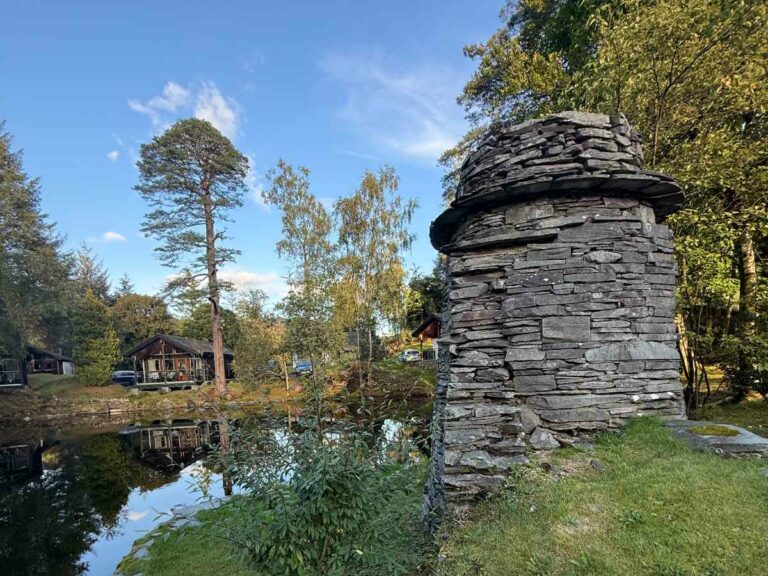 Stacked stone ruins near water with forest cabins and tall trees in background