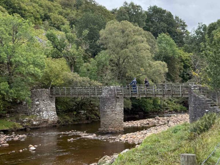 Two hikers crossing historic stone bridge over river in lush Yorkshire Dales woodland