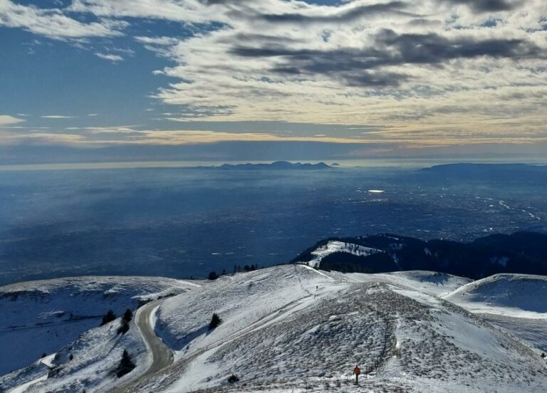 Hiker on snowy Alpine ridge overlooking distant mountains and valley landscape under cloudy sky