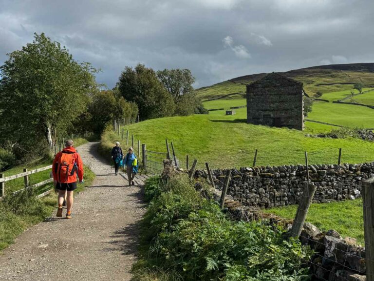 Group of hikers walking along a rural footpath past stone walls and an old barn in green countryside